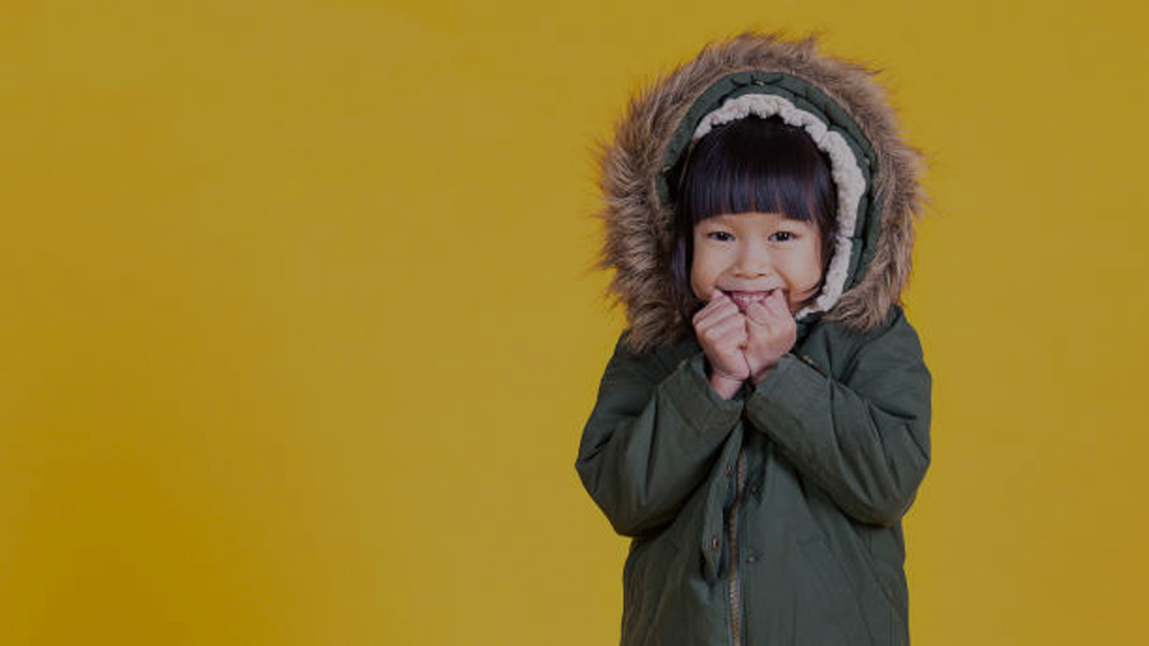 Portrait of young happy little asian girl wearing fur hooded winter coat sweater isolated on yellow background with copy space. Education childhood lifestyle, winter cold banner